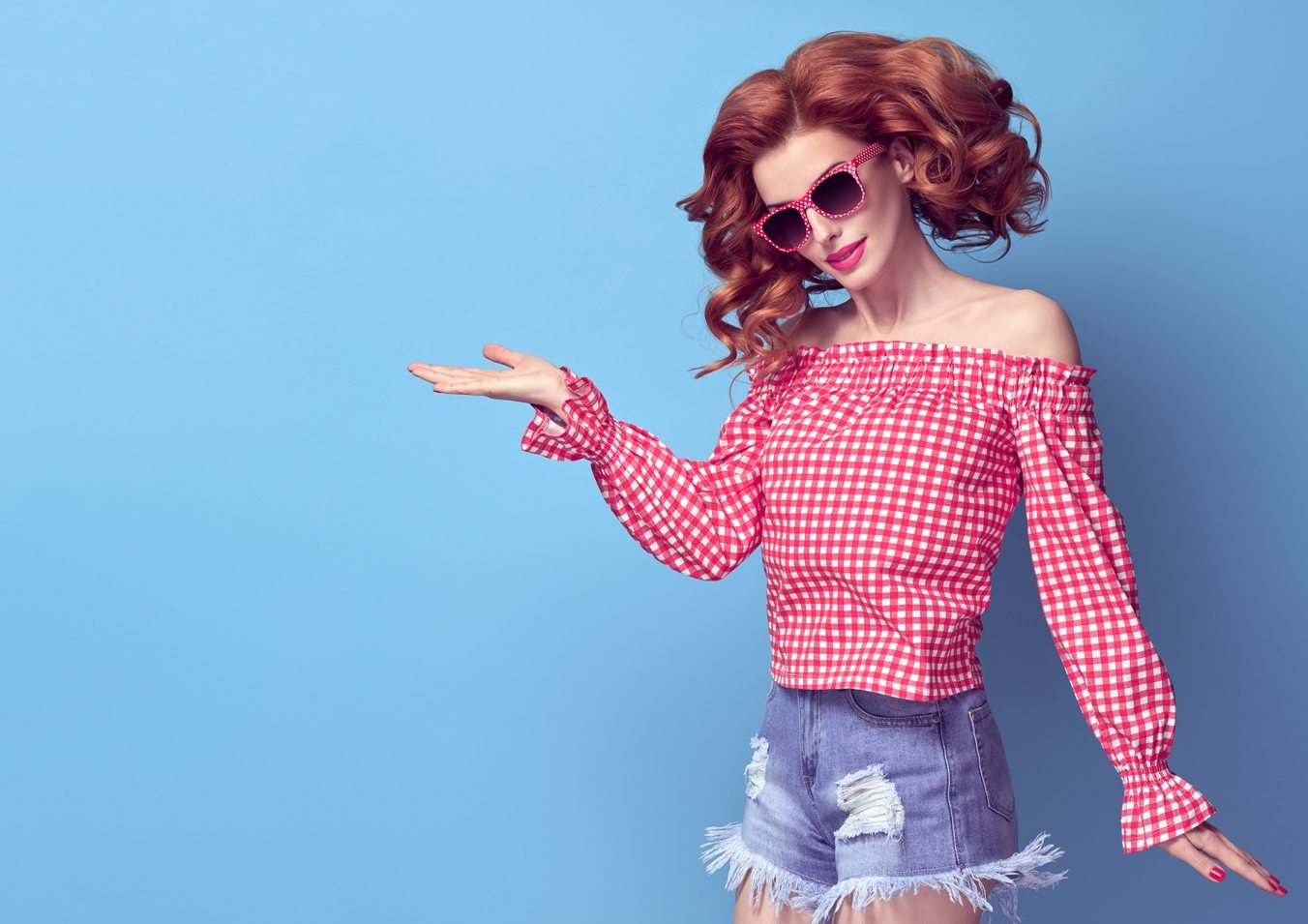 Fashion Pretty PinUp Redhead Girl Smiling Having Fun. Presenting your product. Playful Woman in Pink Summer Bardot Top, Denim shorts, Sunglasses. Trendy Curly hairstyle