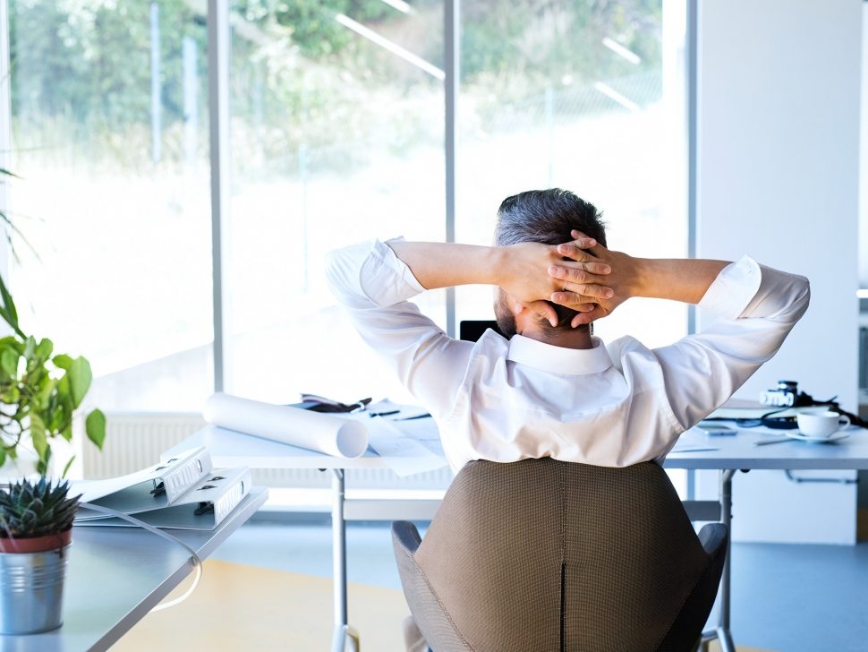 Handsome young businessman in his office, sitting at the desk, stretching arms. Rear view.