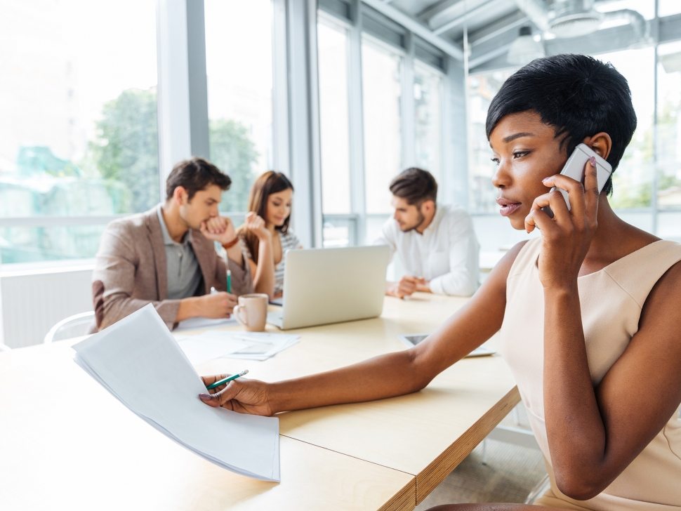 Serious successful young businesswoman with documents sitting and talking on cell phone in office