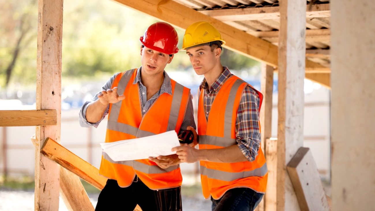 Two men dressed in shirts, orange work vests and helmets explore construction documentation on the building site near the wooden building constructions .