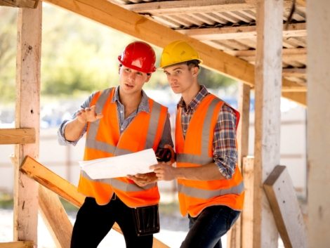 Two men dressed in shirts, orange work vests and helmets explore construction documentation on the building site near the wooden building constructions .