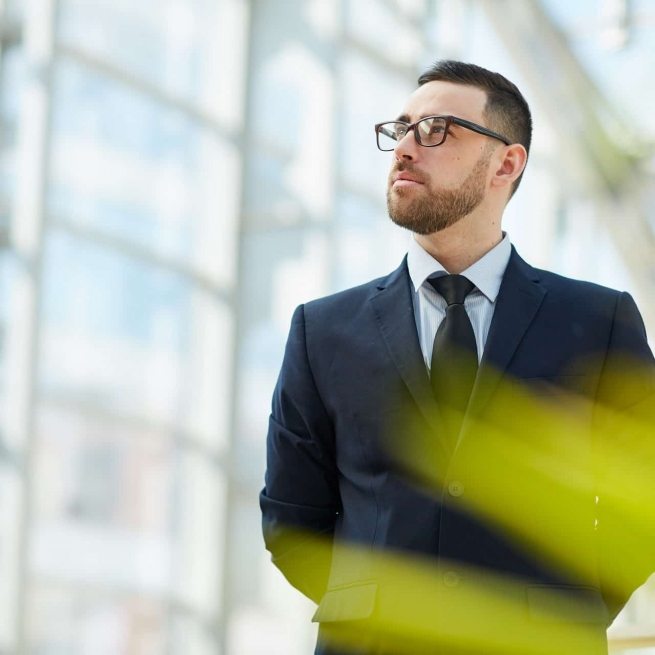 Serious pensive young bearded businessman in eyeglasses being confident in success standing in modern lobby