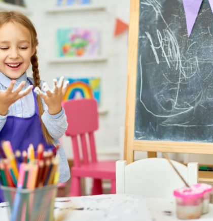 Portrait of cute little girl enjoying art and craft lesson in development school and smiling happily looking at hands covered in paint