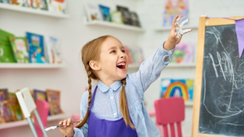 Portrait of adorable little girl enjoying art and craft lesson in development school and smiling happily with hands in paint