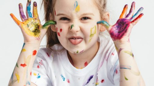 Little girl covered in paint making funny faces. Kindergarten kid. Prankster.