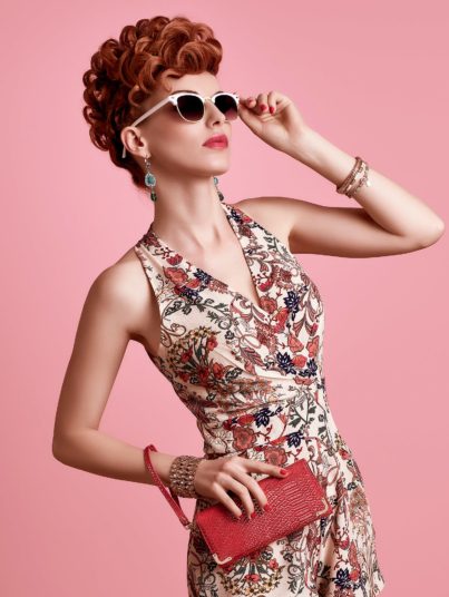Fashion Model in Sexy Jumpsuit. Stylish Mohawk hairstyle, fashion Sunglasses, Summer Floral Outfit. Beauty Redhead Woman in Trendy Summer Dress. Glamour fashion Clutch. Playful Luxury Girl on Pink