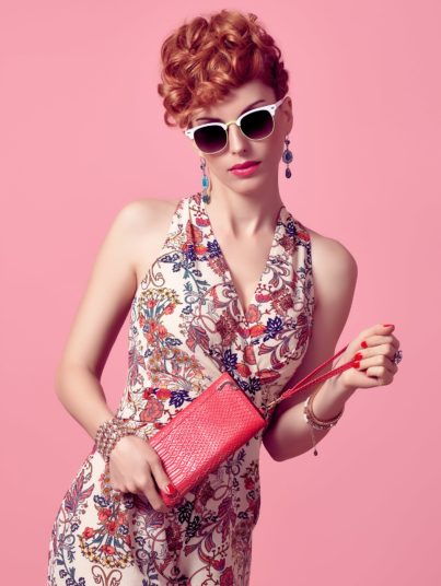 Fashion Redhead Model in Sexy Jumpsuit, woman in Trendy Summer Dress. Stylish Curly hairstyle, fashion Sunglasses, Summer Floral Outfit.Glamour fashion pose.Playful Beauty Girl, Luxury Pink Clutch